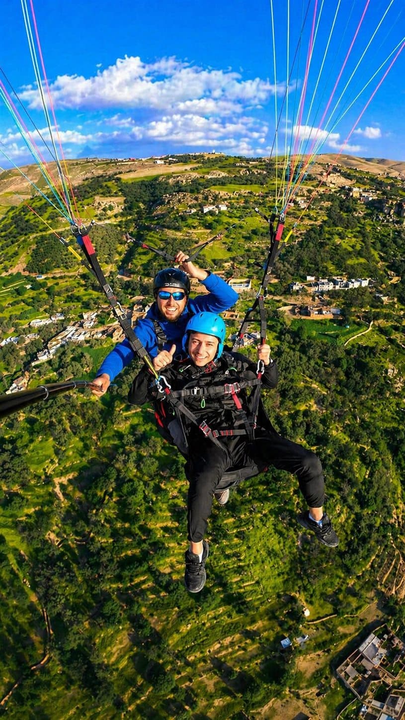 Atlas Paragliding tandem selfie above green foothills with colorful canopy lines.